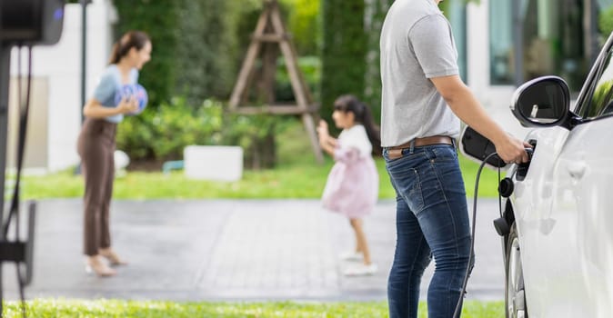 Focus image of progressive man charging electric car from home charging station with blur mother and daughter playing together in the background.