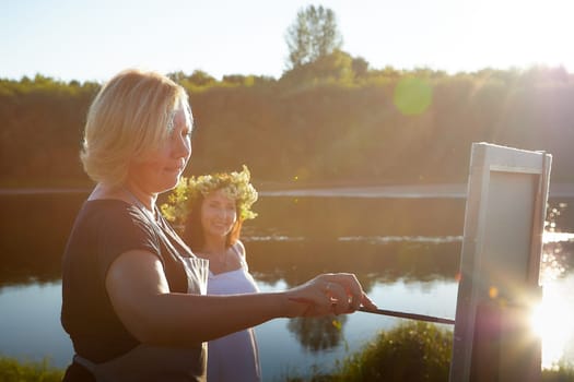 Adult female artist painting picture near water of river or lake in nature. Girl in white sundress and flower wreath for holiday of Ivan Kupala in nature at sunset near artist