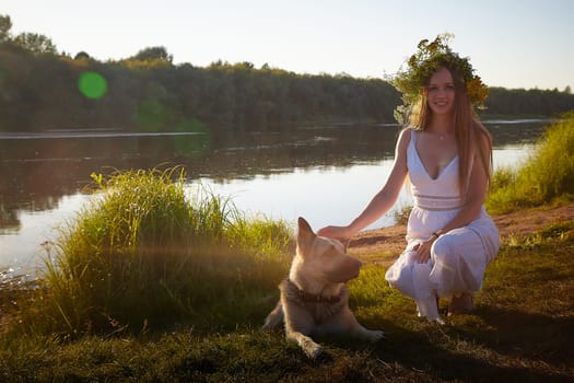 Young brunette girl in a white dress, sundress and wreath of flowers with big shepherd dog in summer on the coast of river or lake in the evening at sunset celebrating pagan holiday of Ivan Kupala