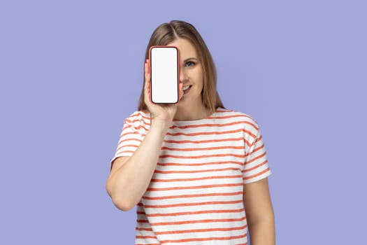 Portrait of cheerful positive blond woman wearing striped T-shirt covering her eye with smart phone with white empty display, copy space for promotion. Indoor studio shot isolated on purple background