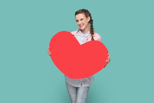 Portrait of flirting positive attractive teenager girl with braids wearing pink jacket standing holding big red heart, winking to camera. Indoor studio shot isolated on green background.