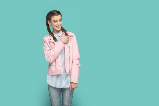 Portrait of smiling positive happy teenager girl with braids wearing pink jacket pointing aside at advertisement area, space for promotion. Indoor studio shot isolated on green background.