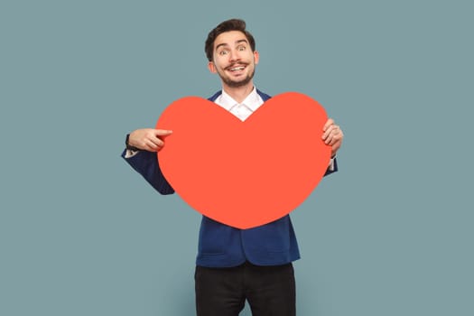 Portrait of handsome man with mustache standing holding pointing at big red heart, smiling toothily to camera, wearing white shirt and jacket. Indoor studio shot isolated on light blue background.
