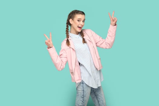 Portrait of excited optimistic cheerful teenager girl with braids wearing pink jacket showing v signs, being happy, being in high spirit. Indoor studio shot isolated on green background.