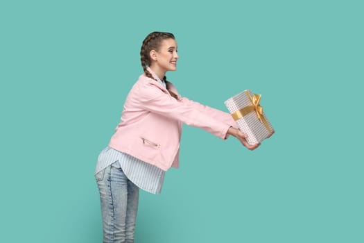 Side view portrait of excited amazed teenager girl with braids wearing pink jacket giving wrapped present box with gold ribbon, congratulating. Indoor studio shot isolated on green background.