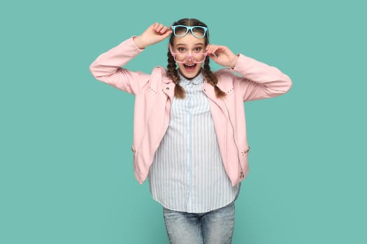 Excited amazed teenager girl with braids wearing pink jacket standing in two colorful optical spectacles, looking at camera with surprised expression. Indoor studio shot isolated on green background.