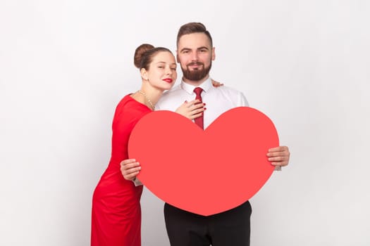 Portrait of pretty pleased loving couple in elegant attires standing together and hugging, man holding big heart, looking at camera. Indoor studio shot isolated on gray background.