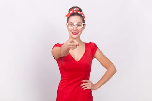 Portrait of positive woman wearing red dress and head band standing with hand on hip, pointing and choosing you, expressing optimism. Indoor studio shot isolated on gray background.