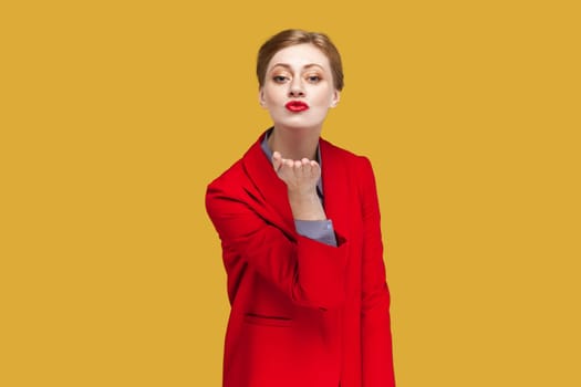 Portrait of romantic woman with red lips standing blowing air kiss over palm, looking at camera, expressing love, wearing red jacket. Indoor studio shot isolated on yellow background.