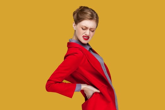 Side view portrait of sad sick unhealthy woman with red lips standing with frowning face, touching her painful back, wearing red jacket. Indoor studio shot isolated on yellow background.