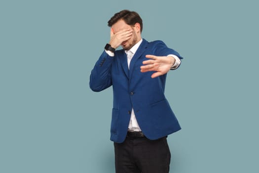 Portrait of man with mustache standing and covering eyes with palm, showing stop gesture, doesn't want to see, wearing official style suit. Indoor studio shot isolated on light blue background.