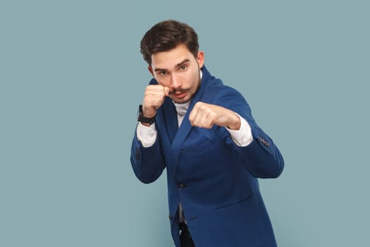 Serious confident man standing with clenched fists, being ready boxing, looking at camera with angry look, wearing white shirt and jacket. Indoor studio shot isolated on light blue background.