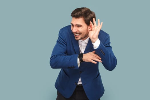 Portrait of handsome man standing with frowning face and hand near ear, trying to listen silent conversation, wearing official style suit. Indoor studio shot isolated on light blue background.