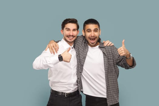 Portrait of two satisfied men friends, standing together and hugging, showing thumbs up, recommend service, good feedback, Indoor studio shot isolated on light blue background.
