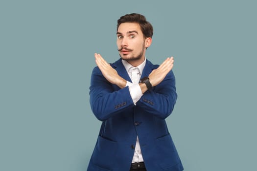 Serious strict man with mustache standing showing x sign, no way gesture, looking at camera with bossy expression, wearing official style suit. Indoor studio shot isolated on light blue background.