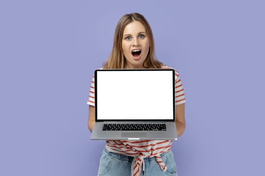 Portrait of shocked amazed blond woman wearing striped T-shirt holding laptop with white empty display with empty space for advertisement. Indoor studio shot isolated on purple background.