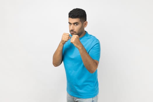 Portrait of confident serious unshaven man wearing blue T- shirt standing with clenched fists, being ready to attack, arguing with somebody. Indoor studio shot isolated on gray background.