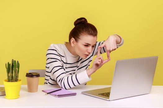 Concentrated woman designer or photographer making photo frame with hands and focusing at laptop screen, cropping picture. Indoor studio studio shot isolated on yellow background.