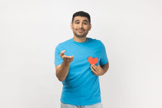 Portrait of falling in love romantic unshaven man wearing blue T- shirt standing giving his heart to his girlfriend, looking at camera, expressing gentle. Indoor studio shot isolated on gray background