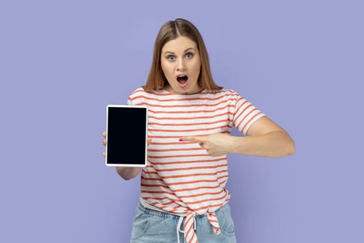 Portrait of amazed shocked surprised blond woman holding pointing at tablet with empty display, mockup screen for your advertisement. Indoor studio shot isolated on purple background.