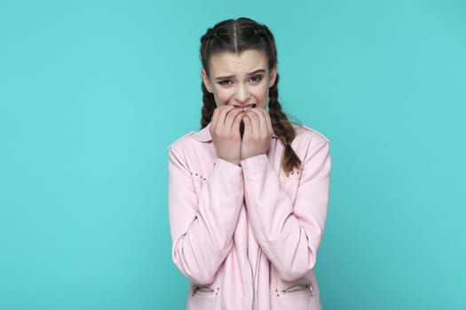 Portrait of nervous stressed teenager girl with braids wearing pink jacket being in deep depression, feels panic attack, biting her fingernails. Indoor studio shot isolated on green background.