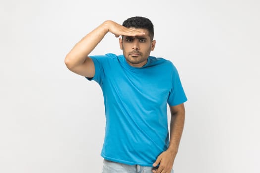 Portrait of unshaven man wearing blue T- shirt standing keeps hand near forehead, looks far away, searches something on horizon. Indoor studio shot isolated on gray background.