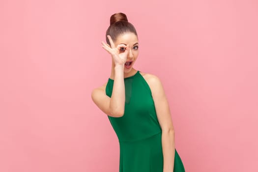 Portrait of funny childish young adult woman standing covering her eye with ok gesture, looking at camera, showing okay sign, wearing green dress. Indoor studio shot isolated on pink background.