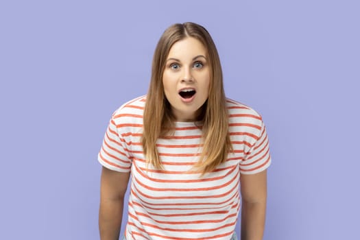 Portrait of blond woman wearing striped T-shirt standing with mouth open in surprise, has shocked expression Indoor studio shot, sees something astonishing. isolated on purple background.