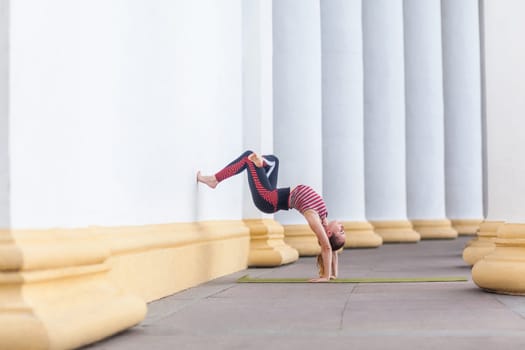 Full length side view of slim attractive athletic sportswoman practicing yoga outdoor, doing wall-assisted handstand pose, wearing stylish sportswear, enjoying acrobatic exercises in open air.