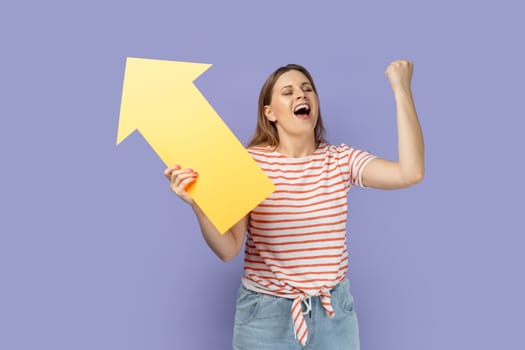 Portrait of extremely happy woman wearing striped T-shirt holding big yellow arrow pointing aside and clenched fist, celebrating victory, increase. Indoor studio shot isolated on purple background.