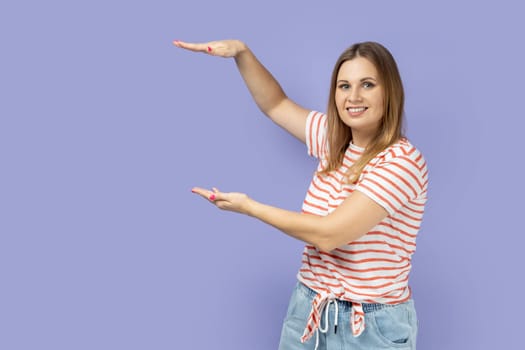 Portrait of satisfied smiling blond woman wearing striped T-shirt presenting area between hands for advertisement, showing huge size. Indoor studio shot isolated on purple background.