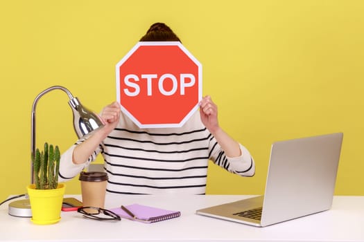 Woman office worker sitting at workplace hiding face behind red stop traffic sign avoiding conflicts, afraid of workplace bullying. Indoor studio studio shot isolated on yellow background.