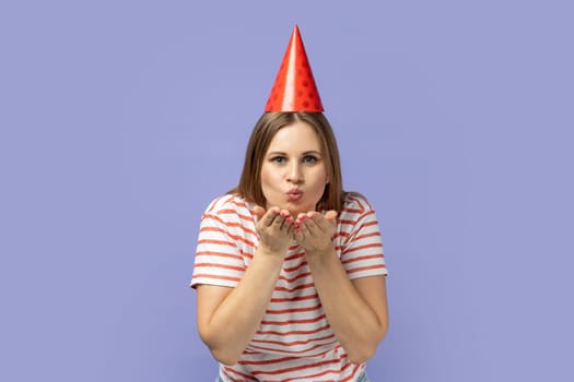 Portrait of extremely happy blond woman wearing striped T-shirt and party cone thanks for the congratulations, sending air kisses to her guests. Indoor studio shot isolated on purple background.