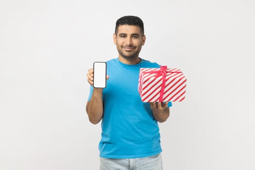 Portrait of cheerful attractive young adult unshaven man wearing blue T- shirt standing with present box and showing cell phone with empty display. Indoor studio shot isolated on gray background.