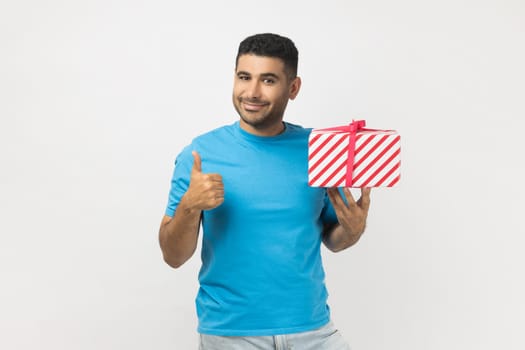 Portrait of attractive joyful unshaven man wearing blue T- shirt standing holding wrapped present box, showing like gesture, thumb up, congratulating. Indoor studio shot isolated on gray background.