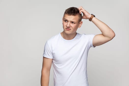 Portrait of thoughtful attractive teenager boy wearing T-shirt standing keeping hand on head, thinking about problem solution, finding answer. Indoor studio shot isolated on gray background.