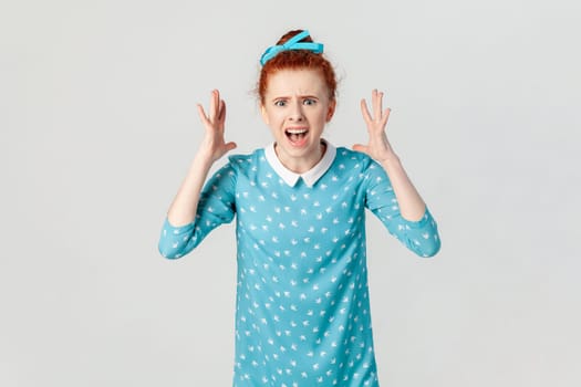 Portrait of angry depressed ginger woman arguing with her husband, screaming with hate and anger, being aggressive, wearing blue dress. Indoor studio shot isolated on gray background.