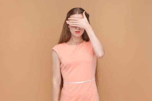 Portrait of serious sad woman with long hair standing and covering her eyes with her palms, doesn't want to see something shame, wearing elegant dress. Indoor studio shot isolated on brown background.