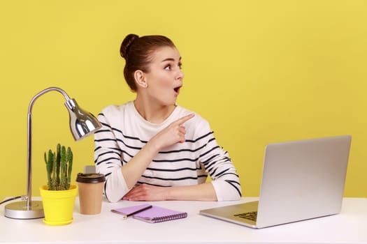 Shocked amazed young woman sitting at office workplace with laptop, looking and pointing aside, showing copy space for promotional text. Indoor studio studio shot isolated on yellow background