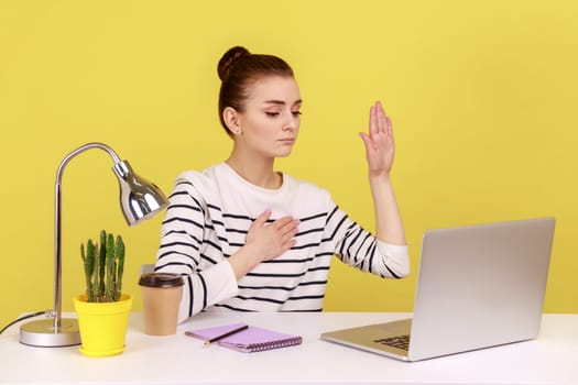 Honest woman sitting at workplace and raising hand to give promise, looking at work place, taking oath with trustworthy faithful expression. Indoor studio studio shot isolated on yellow background.