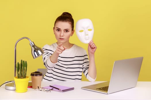 Serious woman office worker holding white face mask and pointing finger at camera, blaming you for hiding identity, duplicity. Indoor studio studio shot isolated on yellow background.