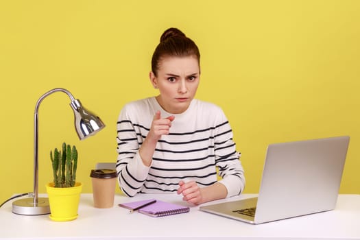 Portrait of serious woman office employee wearing striped shirt sitting on workplace, scolding, saying I have told you, looking at camera. Indoor studio studio shot isolated on yellow background.