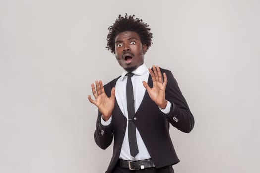 Portrait of scared man with Afro hairstyle stretches hands as shows stop gesture, sees something scary, wearing white shirt and tuxedo. Indoor studio shot isolated on gray background.