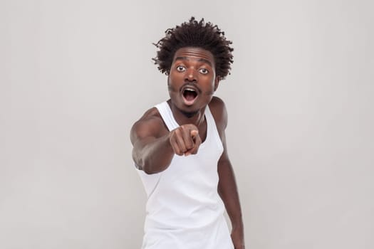 Omg look there. Shocked man with Afro hairstyle points index finger at camera with great surprise, holds breath indicates forward wearing white T-shirt. Indoor studio shot isolated on gray background.
