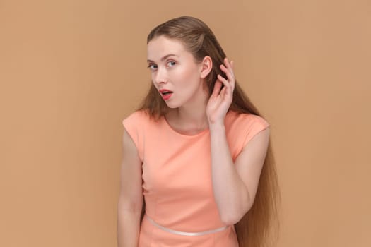 Portrait of attarctive lovely woman with long hair standing with hand near ear, listening to somebody whisper, wearing elegant dress. Indoor studio shot isolated on brown background.