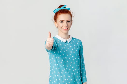 Portrait of satisfied positive optimistic red haired woman with bun hairstyle, standing showing like gesture, recommend, wearing blue dress. Indoor studio shot isolated on gray background.
