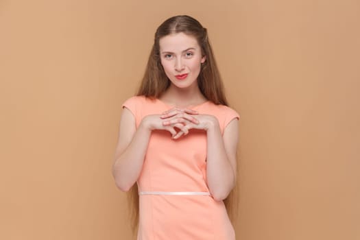 Portrait of cunning sly attractive woman with long hair standing with playful bitchy facial expression, planning devil prank, wearing elegant dress. Indoor studio shot isolated on brown background.