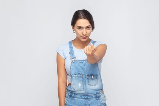 Come here. Portrait of attractive woman wearing denim overalls gesturing come to me, beckoning with finger, inviting for confidential talk. Indoor studio shot isolated on gray background.