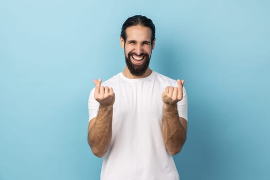 Portrait of smiling happy positive man with beard wearing white T-shirt hair makes korean like sign mini hear gesture snaps fingers. Indoor studio shot isolated on blue background.
