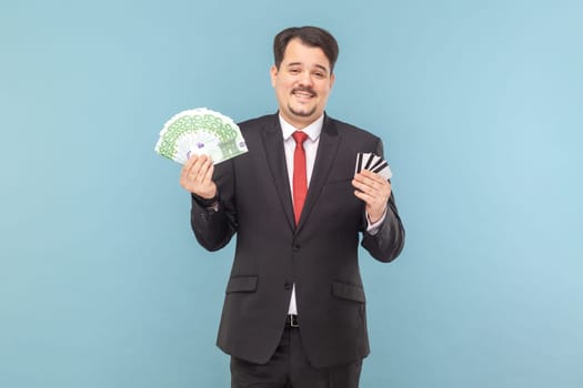 Satisfied rich man with mustache standing holding euro banknotes and credit cards in hands, being happy, wearing black suit with red tie. Indoor studio shot isolated on light blue background.